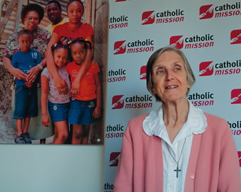 Sister Anne Quinn SMSM before a poster featuring Sister Teresia SMSM and a Jamaican  Family.