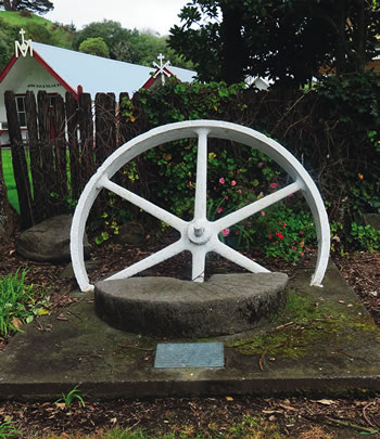 The plaque reads: This wheel and stone were once part of the Pukekaraka flour mill erected about 1854 under the direction of Father Comte s.m. The mill stood on the bank of the Waitohu River on property now owned by Mr Ray Taylor who donated these remains for erection on the site of the Pukekaraka Mission. Otaki Historical Society Inc. 1985